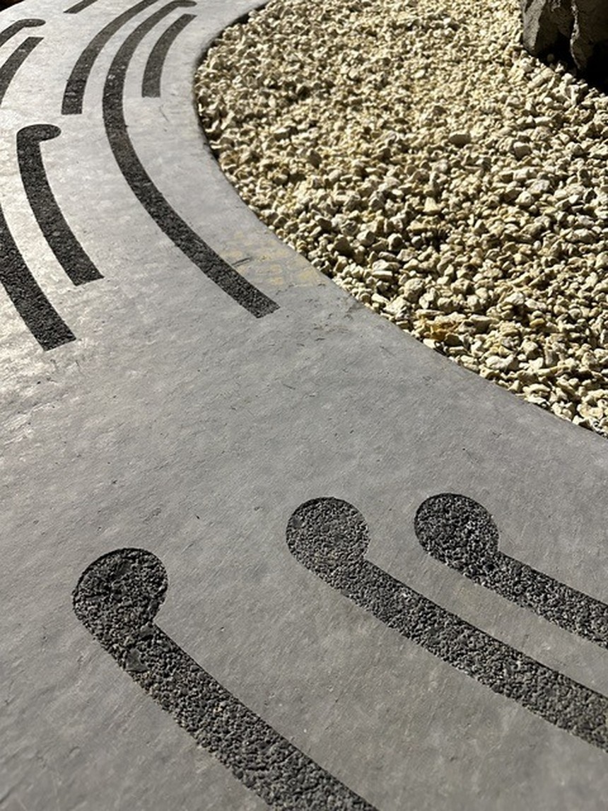 Surface detail: black textured line channels curving across a pale cast-concrete field beside river stones